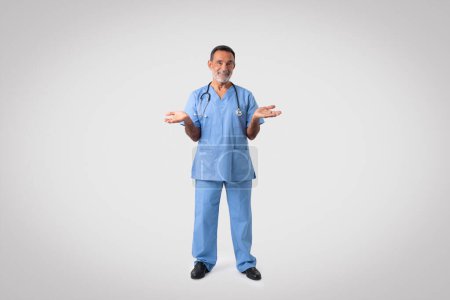 Photo for Happy caucasian senior doctor surgeon in blue uniform hold free space on hands, isolated on gray background, studio. Choice for medicine, ad and offer, health care, professional work and advice - Royalty Free Image