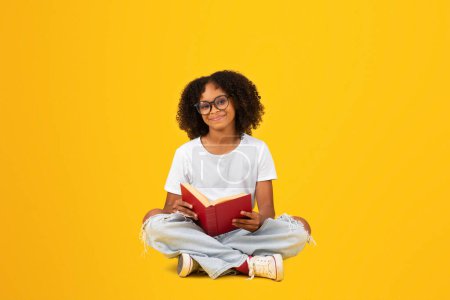 Photo for Cheerful smart curly teenager black schoolgirl in white t-shirt and glasses reads book, isolated on yellow background, studio. Study, knowledge, hobbies, education and homework, exam preparation - Royalty Free Image