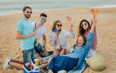 Foto de Sonriendo millennial europeo y árabe personas en casual divertirse en gafas, disfrutar de la fiesta en la playa, beber cerveza en fin de semana, al aire libre. Picnic con amigos, vacaciones juntos en verano - Imagen libre de derechos