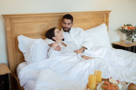 Photo for Honeymoon Pleasure. Loving Couple Hugging Each Other During Their Breakfast In Bed At Hotel Bedroom Indoors. Young Husband And Wife Cuddling At Luxury Suite In The Morning - Royalty Free Image
