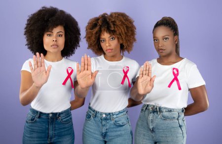 Photo for Oncology Awareness. Three Determined Black Women In T-Shirts With Pink Breast Cancer Ribbons Gesturing Stop Standing Over Purple Background, Looking At Camera. Studio Shot - Royalty Free Image