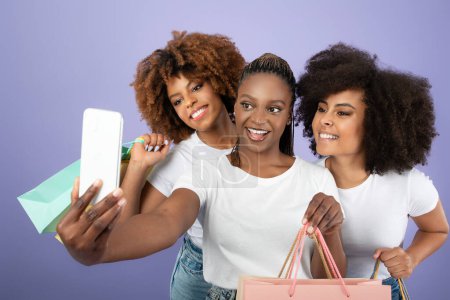 Téléchargez les photos : Le commerce électronique. Trois joyeuses femmes noires faisant du shopping et s'amusant à faire du selfie sur un téléphone portable, posant des sacs à provisions en papier sur fond violet. Mode et concept de style. Studio Shot - en image libre de droit