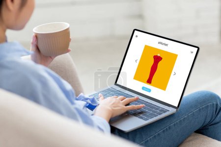 Photo for Lady with coffee cup sitting on sofa, using tlaptop pc with fashion store on screen, choosing dress and ordering clothes at home, enjoy online shopping - Royalty Free Image
