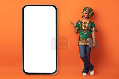 Foto de Sonriente mujer negra atractiva positiva en traje africano con el ordenador portátil en la mano apuntando al teléfono grande con pantalla blanca, mostrando buen sitio web o aplicación, fondo naranja, espacio de copia, maqueta - Imagen libre de derechos