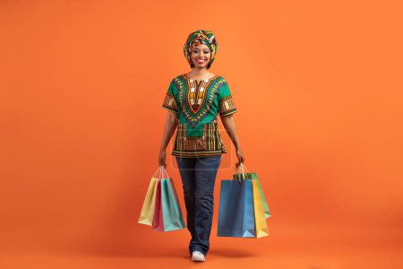 Photo for Black friday, sales season, shopping concept. Happy cheerful attractive young black lady in traditional african costume walking with her purchases, isolated on orange studio background, copy space - Royalty Free Image