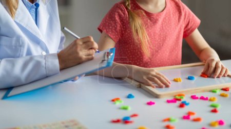 Photo for Unrecognizable little girl making words from colorful plastic letters during meeting with psychologist, child development specialist exercising with female kid and taking notes to clipboard, cropped - Royalty Free Image
