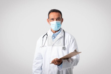 Photo for Serious caucasian mature man doctor in white coat and protective mask, isolated on gray background, studio. Disease treatment, diagnostics, health care, ad and offer, medicine service - Royalty Free Image