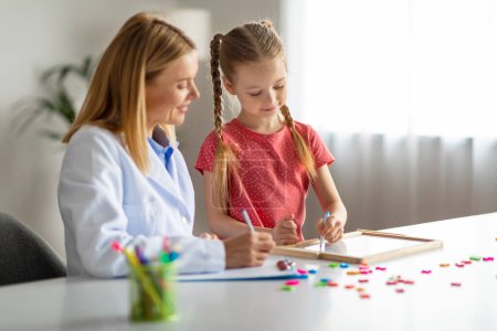 Photo for Cute Little Girl Having Therapy Meeting With Professional Child Psychologist Woman, Smiling Female Kid Sitting At Table And Writing On Drawing Board, Learning Letters, Treating Dyslexia, Copy Space - Royalty Free Image