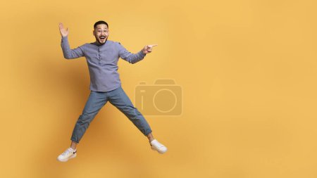 Téléchargez les photos : Positif jeune homme asiatique montrant l'espace de copie tout en sautant dans l'air, heureux Millennial Guy s'amuser sur fond jaune studio, Démonstration endroit gratuit pour la conception de la publicité, Panorama - en image libre de droit