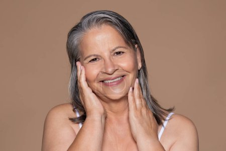 Photo for Happy european elderly woman with gray hair touches face with hands, isolated on beige studio background, close up. Spa procedures result, beauty care, anti-aging, anti-wrinkle treatment - Royalty Free Image