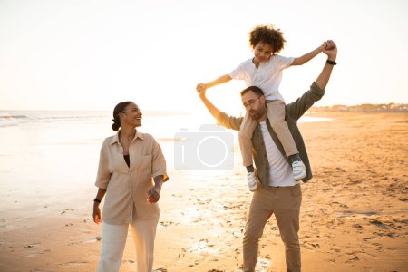 Photo for Happy multiracial family of three walking by seaside in the evening, father carrying son on shoulders while walking together outside, enjoying warm summer day - Royalty Free Image