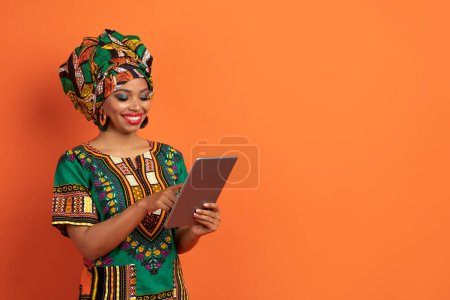 Photo for Great mobile app. Stylish cheerful happy beautiful young arican american lady wearing bright traditional costume using modern digital tablet on orange studio background, copy space - Royalty Free Image