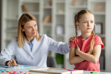 Psychotherapeutin tröstet aufgebrachtes kleines Mädchen während Therapiesitzung im Büro, Spezialistin für kindliche Entwicklung versucht Patientin zu beruhigen, beleidigtes Kind steht mit verschränkten Armen