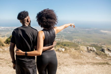 Téléchargez les photos : Femme afro-américaine embrassant l'homme profitant de la pause d'entraînement, pointant du doigt, debout sur des rochers à l'océan, à l'extérieur, vue de dos, espace libre. Mode de vie actif et sport - en image libre de droit