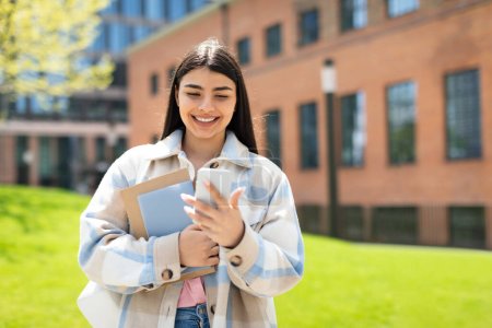 Téléchargez les photos : Étudiante hispanique joyeuse avec smartphone et classeurs debout à l'extérieur, marchant sur le campus de l'université après les cours. Heureux chat féminin avec des amis et souriant - en image libre de droit