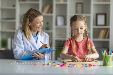 Photo for Kids Psychology Concept. Smiling Psychotherapist Lady Taking Notes During Session With Little Girl, Cute Female Child Making Word With Colorful Letters On Table, Enjoying Development Games, Closeup - Royalty Free Image