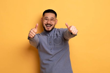 Photo for All Great. Positive Young Asian Man Showing Thumbs Up At Camera, Cheerful Millennial Guy In Eyeglasses Gesturing Sign Of Approval, Recommending Something While Standing Over Yellow Background - Royalty Free Image