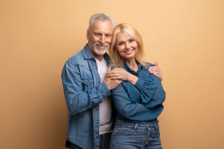 Photo for Handsome loving elderly man husband hugging his beautiful blonde wife, senior couple in casual denim outfits cuddling on beige studio background, enjoying time together, copy space - Royalty Free Image