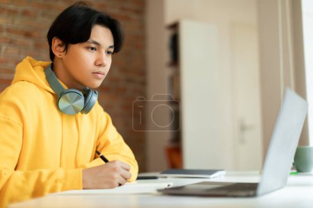 Téléchargez les photos : Concept d'apprentissage en ligne. Adolescent asiatique intelligent gars regardant l'écriture d'ordinateur portable dans un ordinateur portable pendant la leçon Web à la maison. Étudiant occupé participant à une conférence en ligne assis au bureau intérieur, vue latérale - en image libre de droit