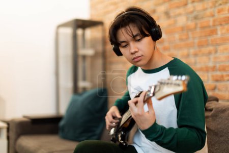 Photo for Musical Creativity. Portrait of Young Chinese Teen Guy Playing Electric Guitar with Earphones On, Learning To Play At Home, Sitting On Sofa. Talented Musician Engaged In Music Lesson. Hobby And Art - Royalty Free Image