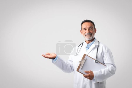 Photo for Positive caucasian mature man doctor in white coat holds empty space in his hand, isolated on gray background, studio. Professional advice, health care, ad and offer, medical service - Royalty Free Image