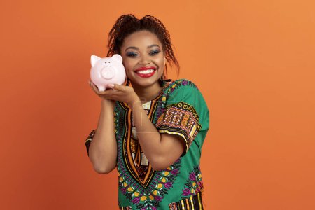 Foto de Educación financiera, concepto de ahorro. Emocionado feliz linda mujer negra bastante joven en traje africano tradicional celebración de alcancía junto a su cara y sonriente, fondo naranja - Imagen libre de derechos