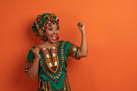 Foto de Emocional emocionado atractiva joven mujer negra en traje africano gesto en fondo de estudio naranja, dama ganadora apretando puños y exclamando, mirando espacio de copia - Imagen libre de derechos