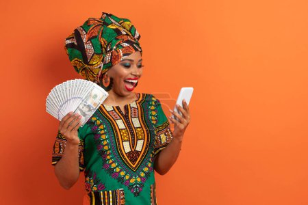 Photo for Emotional happy millennial african american lady in traditional costume with money cash dollar banknotes and smartphone in her hands exclaiming, isolated on orange background, copy space - Royalty Free Image