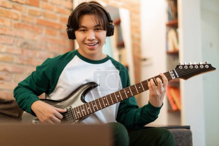 Photo for Excited Young Asian Guy Practicing Guitar Chords Looking At Laptop Indoor. Teenager Boy Sitting With Electric Guitar Wearing Wireless Headphones, Enjoying His Musical Hobby At Home - Royalty Free Image