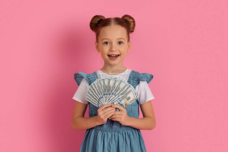 Photo for Cute Little Girl Holding Dollar Cash And Smiling At Camera, Happy Preteen Female Child Showing Money Banknotes, Enjoying Savings And Economy While Standing On Pink Background, Copy Space - Royalty Free Image