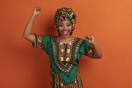 Foto de Super feliz emocional hermosa joven afroamericana dama vistiendo traje tradicional y maquillaje brillante gesto sobre fondo naranja, levantando los puños y gritando, celebrando la victoria del éxito - Imagen libre de derechos