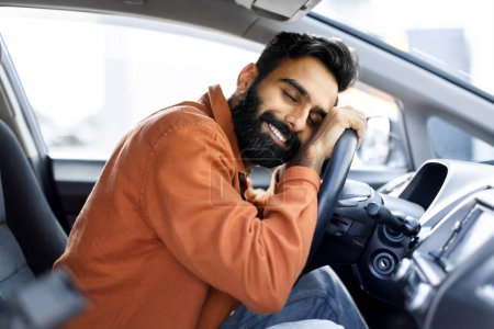 Foto de El sueño se hace realidad. Hombre indio feliz abrazando el volante de su nuevo auto sentado en el asiento de los conductores. Alegre barbudo chico celebrando propia compra de automóviles sentado en el interior del vehículo - Imagen libre de derechos