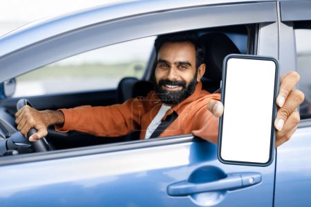 Foto de Alegre chico árabe mostrando su teléfono móvil pantalla en blanco a través de la ventana abierta del coche, conducción de vehículos y sonriendo a la cámara, enfoque selectivo. Driver Man Publicidad Aplicación de navegación - Imagen libre de derechos