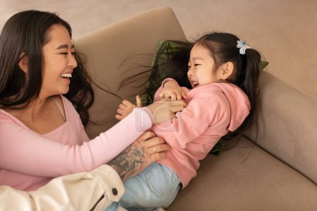 Foto de Bono familiar y diversión. Alegre mujer china cosquillas a su hijita y riendo, disfrutando de tiempo de juego juntos en el sofá en casa. Padres que se unen con Kid el fin de semana. Cultivado - Imagen libre de derechos
