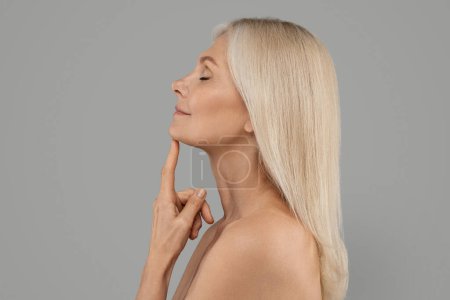 Photo for Anti-Aging Skincare. Side View Shot Of Beautiful Mature Female With Flawless Skin Touching Chin, Attractive Elderly Woman With Bare Shoulders Posing Over Grey Studio Background, Copy Space - Royalty Free Image