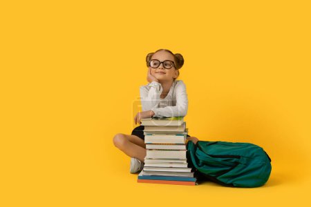 Téléchargez les photos : Mignon élémentaire fille assise à livres empilent sur fond jaune, souriant Preteen enfant féminin avec sac à dos posant dans le studio, heureux élève enfant regardant à côté de l'espace de copie - en image libre de droit