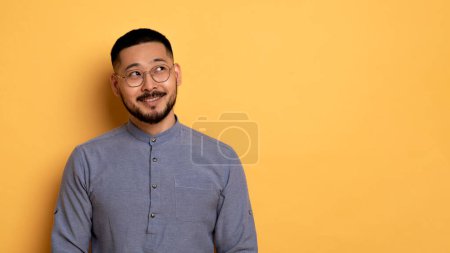 Photo for Pensive Young Asian Man Looking Aside At Copy Space And Smiling, Dreamy Cheerful Millennial Guy Thinking About Interesting Offer, Standing Over Yellow Background In Studio, Panorama - Royalty Free Image