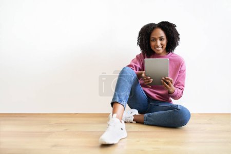 Téléchargez les photos : Joyeux millénariste femme noire assise sur le sol, bavardant sur la tablette dans le salon intérieur sur fond de mur blanc. Étude, application de travail à la maison, chat, blog et réseaux sociaux - en image libre de droit