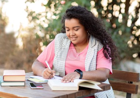 Photo for College Education. Latin Student Woman Studying Outdoors, Taking Notes While Reading Book Sitting At Table In Park. Young Lady Learner Doing Homework, Working On Educational Project Outside - Royalty Free Image