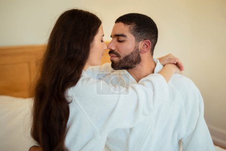 Photo for Love and Relationship. Young Spouses Hugging In Bed Wearing Bathrobes At Hotel Bedroom Indoors. Lovers bonding Sharing Intimate Moments Of Tenderness And Romance. Side View - Royalty Free Image