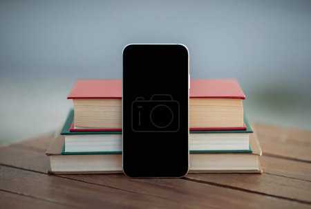 Mobile Anwendung. Smartphone mit leerem schwarzen Bildschirm, der neben einem Stapel Bücher auf einem Tisch im Freien steht. Mockup für Bildungs-App-Werbung mit Phone Gadget. Technologie, E-Learning