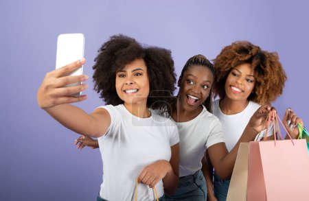 Foto de Amigos de compras. Emocionado trío de damas negras haciendo selfie en el teléfono inteligente, divirtiéndose juntos mientras sostienen bolsas de compras en el estudio sobre fondo púrpura. Oferta de comercio electrónico Anuncio - Imagen libre de derechos