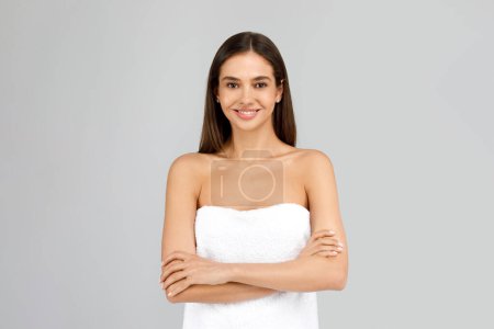 Photo for Beauty concept. Beautiful european young woman wrapped in bath towel posing with folded arms and smiling at camera isolated over grey background, studio shot - Royalty Free Image