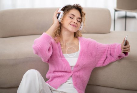 Téléchargez les photos : Belle jeune femme dans les écouteurs sans fil écoutant de la musique avec les yeux fermés à la maison, souriant Millennial Femme bénéficiant de sa playlist préférée sur Smartphone, se détendre sur le sol dans le salon - en image libre de droit