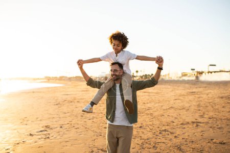 Photo for African american boy sitting on fathers shoulders, family walking on beach by seaside, spending evening together outside, having fun and fooling - Royalty Free Image