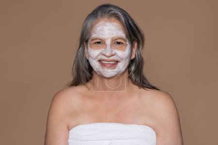 Téléchargez les photos : Glad européenne senior femme aux cheveux gris en serviette profiter de la procédure de spa, masque facial, isolé sur fond studio beige. Beauté, soins de la peau, cosmétiques et traitement hydratant anti-âge - en image libre de droit