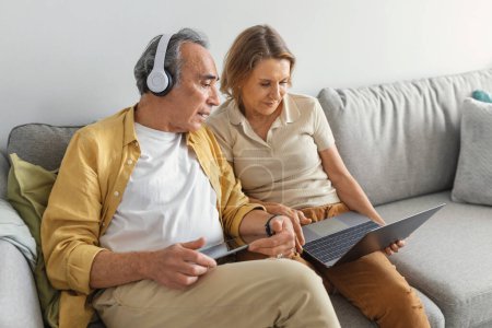 Téléchargez les photos : Grands-parents modernes, homme et femme âgés utilisant un ordinateur portable et une tablette, Internet de surf, mâle portant un casque sans fil, assis sur le canapé à la maison, espace libre - en image libre de droit