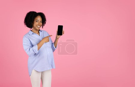 Foto de Feliz milenario negro rizado embarazada dama con el dedo del vientre en el teléfono con la pantalla en blanco, aislado en el fondo del estudio rosa. Recomendación, aplicación para esperar bebé, blog en la maternidad futura - Imagen libre de derechos