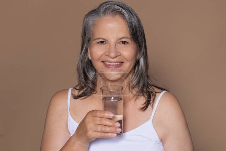 Photo for Happy european senior grey-haired woman take glass of water, enjoy morning routine, isolated on beige studio background, copy space. Health, body and beauty care, diet at menopause - Royalty Free Image
