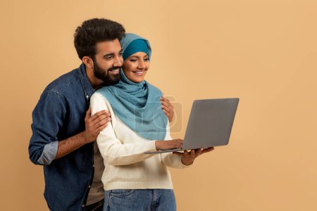 Téléchargez les photos : Jeune couple musulman utilisant ordinateur portable ensemble tout en se tenant sur fond de studio beige, romantique jeune homme arabe et femme dans hijab shopping en ligne ou faire un appel vidéo, espace libre - en image libre de droit